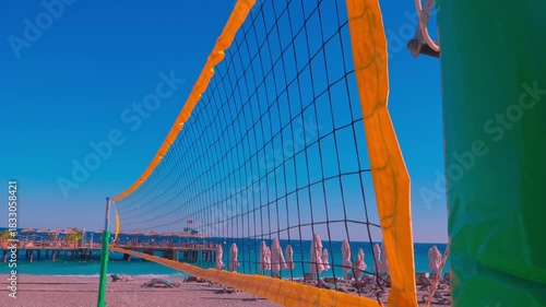 An empty volleyball court on a resort beach comes alive in the breeze, where the net gently sways, creating an atmosphere of tranquil anticipation. Golden sand lapped by the nearby sea and the gentle 