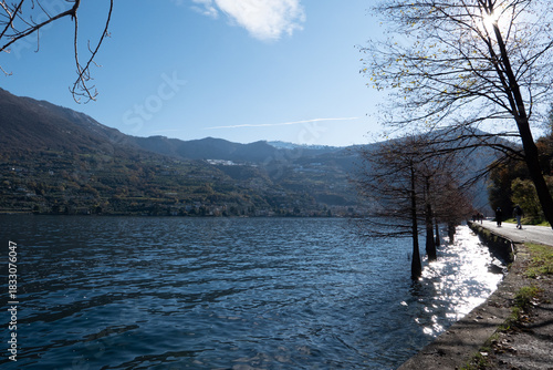 Winter light illuminates the lakeside walkway on Lake Iseo, with bare trees, gentle reflections on the water, and a calm, atmospheric scene capturing the quiet beauty of the season.