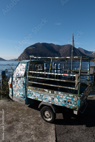 Colorful Piaggio Ape covered in stickers parked by Lake Iseo, creating a quirky and vibrant scene with mountains, calm water, and a playful Italian atmosphere.