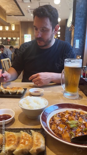 Caucasian man eating gyoza, mapo tofu, rice and beer with chopsticks in a Japanese restaurant