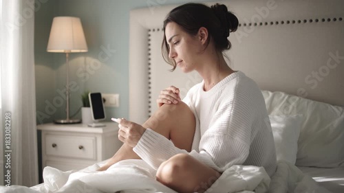 Young woman sitting on bed, smiling while talking on phone, holding pregnancy test, cozy bedroom with soft lighting and white bedding, conveying joy and anticipation of motherhood