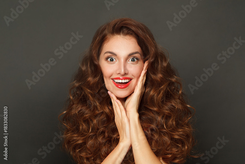 Joyful surprised woman with curly hairstyle and makeup holding her hand with her face on black background
