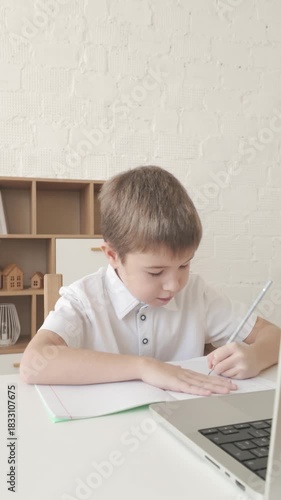 Schoolboy studies during online lesson on laptop, writing task in notebook at desk. Vertical video for education, e-learning and school themes