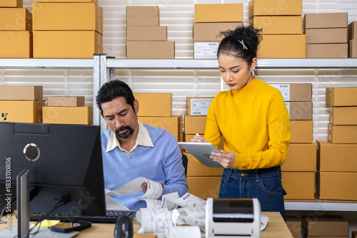 Small business couple analyzing printed labels, recording shipment updates by computer technology, coordinating inventory tasks, e-commerce operations with teamwork, and organizing warehouse planning