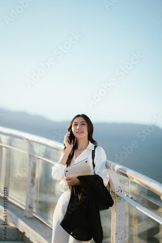 An Asian woman walking outdoors while talking on the phone, carrying a laptop and jacket, portraying a confident Asian businesswoman in a bright, modern setting.