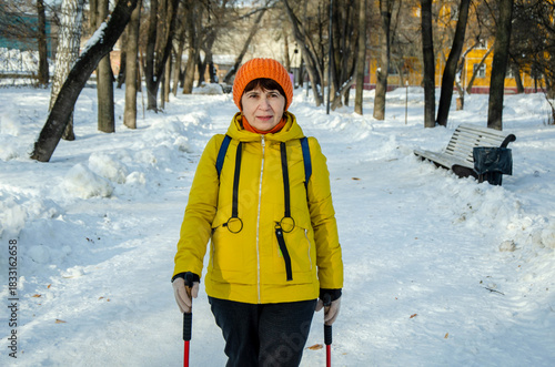 Senior woman in yellow coat doing Nordic walking along snowy path in city park
