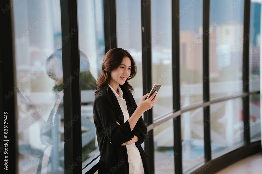 Fototapeta premium A professional woman standing by large windows, smiling while working on a laptop in a bright modern office environment.