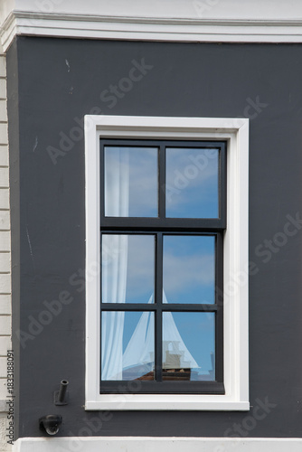 Minimal exterior wall with a black framed window and white trim. Blue sky reflection with a sailboat in the glass and soft curtains inside. Vlaardingen, Netherlands.