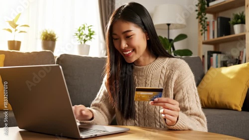 happy young asian woman sitting on sofa, holding credit card and using laptop. online shopping and internet banking payment. e-commerce, consumerism concept. home finance management.