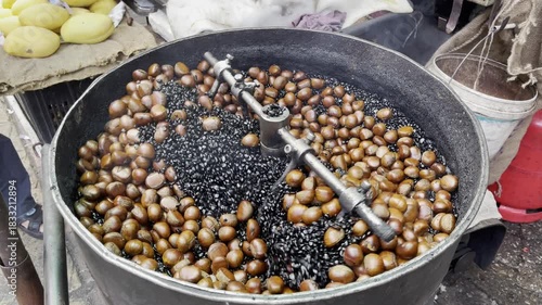 Chestnuts roast in a rotating drum filled with hot black stones at a bustling street market in Kuala Lumpur, releasing rich aroma and showcasing authentic traditional snack preparation