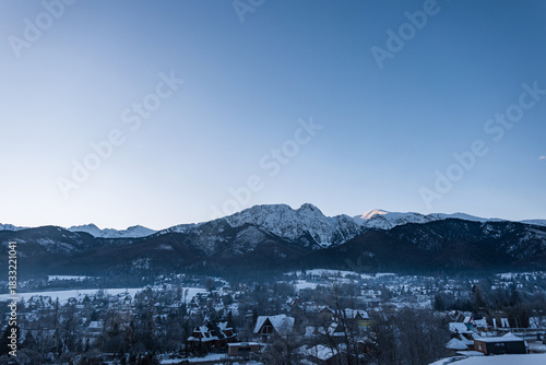 Fototapeta Naklejka Na Ścianę i Meble -  Panoramic view of the misty, snow-covered resort town of Zakopane nestled beneath the towering, rocky peaks of the Tatra Mountains, Poland. Clear blue sky with sun. Winter holidays