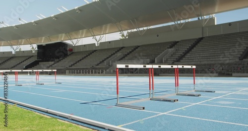 Showing set of red-and-white hurdles crossing blue running track at outdoor stadium, with canopy
