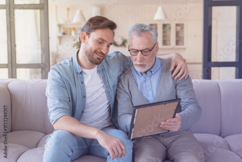 Foto Caring loving adult son hugging embracing his old elderly senior father while he is showing his family photograph photo at home, telling stories of his youth