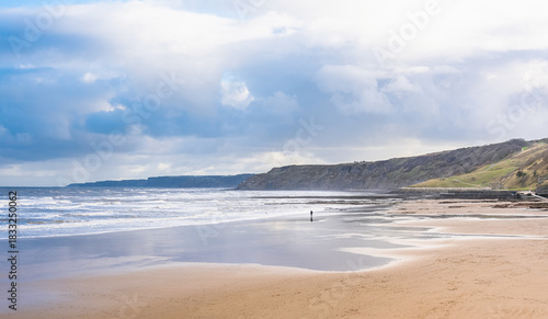 A sandy beach with a rough sea and headlands. A cloudy sky is above.