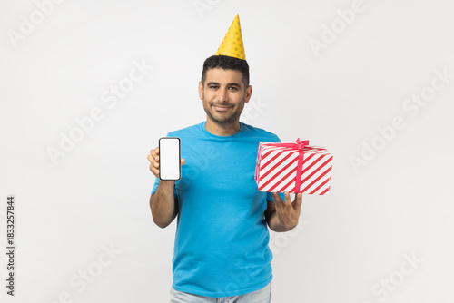 Man wearing yellow party cone holding present box and showing cell phone with empty display.