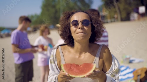 A young woman enjoying a slice of fresh watermelon on the beach while her friends relax and have fun in the background.