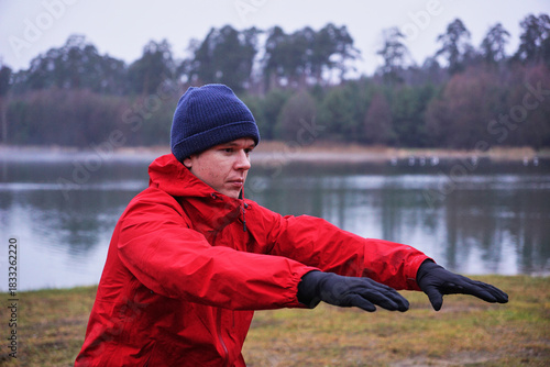 A young athlete is warming up before a workout in a park by a lake. A man is doing squats