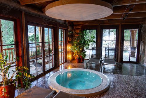Indoor jacuzzi in a warm wooden cabin hotel with large windows overlooking snowy forest .
