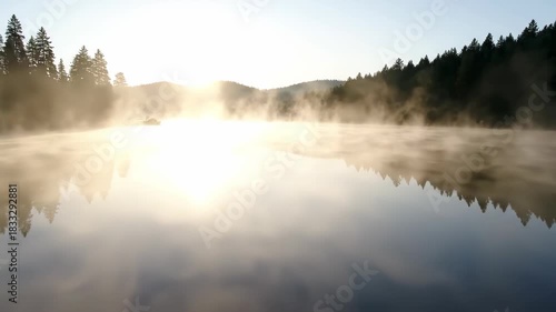 Golden sunrise light glistens on a serene lake, ethereal mist rising over forest trees in early morning light