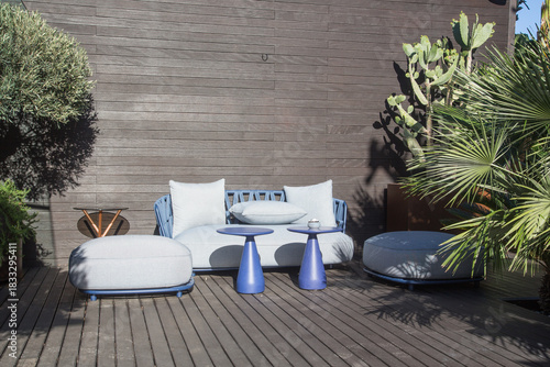 Bar with modern and cozy fabric lounge furniture put on a terrace with wooden pavement surrounded by lush growing palm trees, succulents and green plants