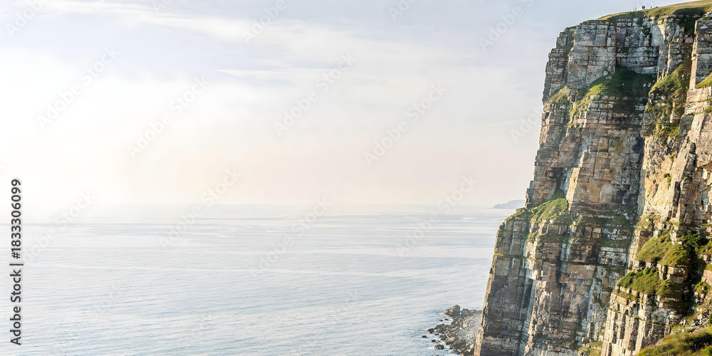 Fototapeta premium Massive, vertical limestone cliff covered in green growth, set against endless ocean and bright, hazy sky.