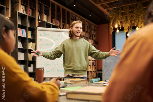 Young man standing in front of whiteboard teaching diverse group of students in library classroom, gesturing with arms open while explaining lesson content