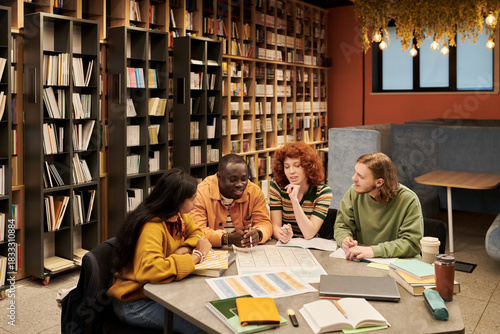 Group of multiethnic young people sitting at table discussing documents and studying together in modern library setting, bookshelves filled with books in background