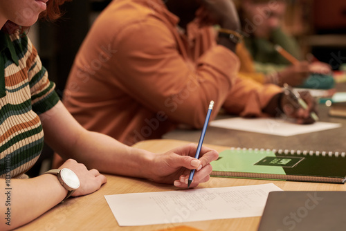 Young woman writing on paper at desk beside Black young man taking notes in classroom setting, both focused on academic work, notebooks and pencils