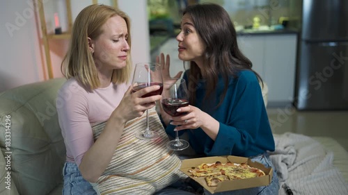 Two close friends are sitting together at home, sharing pizza in a cozy living room. One woman is visibly sad and on the verge of tears, while the other gently comforts her with empathy and support.