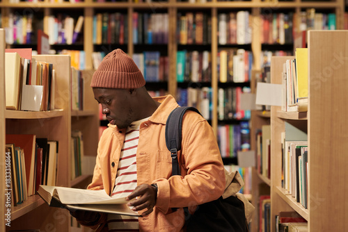 Young Black man standing between bookshelves reading large book, wearing backpack, focusing on pages, surrounded by shelves filled with books in library setting
