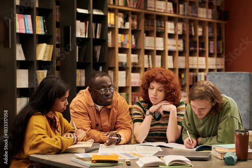 Group of diverse young people studying together at table in library, multiethnic men and women collaborating on academic project, reading books, taking notes, discussing assignments