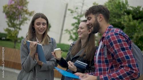 Three students sit together on a college campus, holding books and chatting casually. They smile and enjoy a relaxed moment outdoors as they talk about their studies.