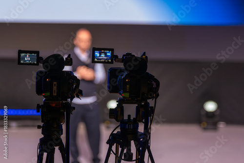 Cameras set up focus on blurred presenter delivering talk while audience unseen. Behind scenes shot highlights media production, stage lights, filming setup for corporate events and interviews