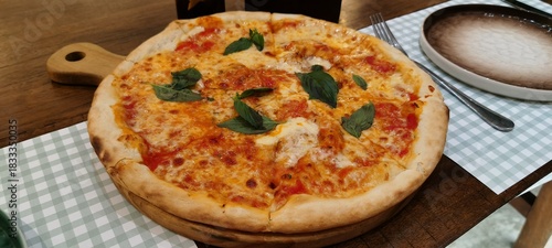 A delicious, freshly baked Margherita pizza with a golden crust, melted mozzarella cheese, bright red tomato sauce, and fresh basil leaves, served on a wooden board at a restaurant table.