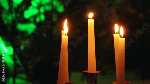 Five tall wax candles burn gently in warm orange hues, set against a softly focused green and dark background, creating a cozy and intimate atmosphere.