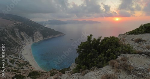 Breathtaking panorama of iconic Myrtos beach in the Greek island Kefalonia, with turquoise waters of Ionian Sea surrounded by steep cliffs, under dramatic sunset red sky, stunning Mediterranean view