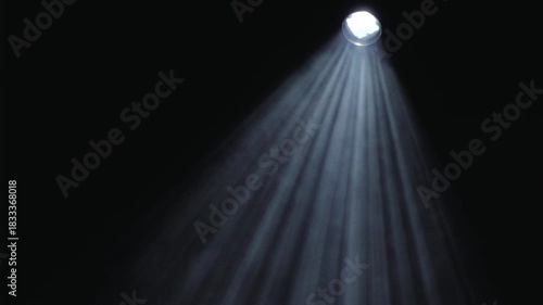 A bright overhead spotlight projects focused beams through darkness, forming visible rays of light in a smoky or dusty environment with ample