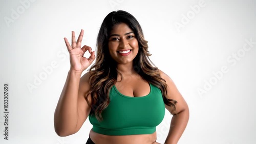 smiling indian plus size woman in green sportswear showing ok sign. body positive female posing on white background. fitness, approval and agreement concept.