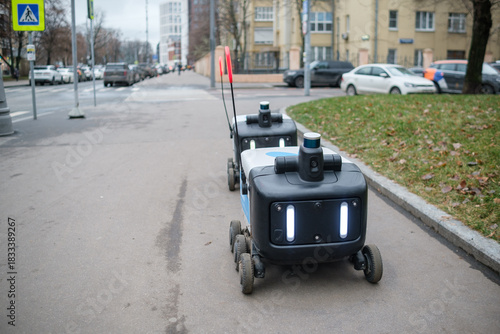 Fototapeta Naklejka Na Ścianę i Meble -  Delivery Robots Navigate Urban Sidewalk in City During Cloudy Weather and Afternoon Hours