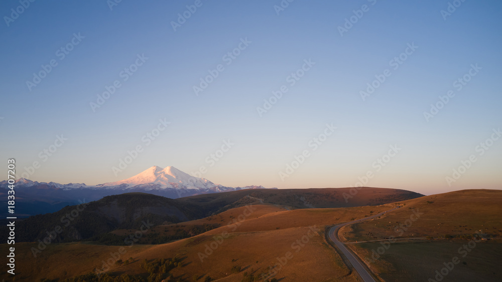 Fototapeta premium Autumn mountain landscape. A winding road across a mountain plateau with a snow-capped volcano in the distance. Drone footage.