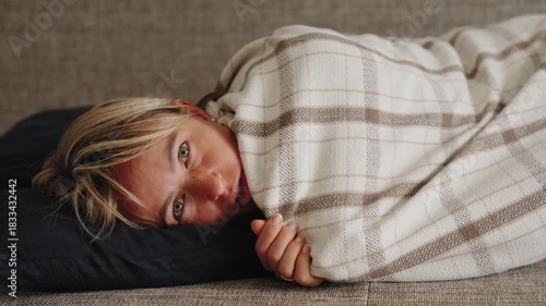 Young woman lies curled on a sofa in red plaid pajamas, holding her lower abdomen in visible discomfort. The TV remote is nearby. Concept of menstrual pain and rest during periods. Natural light, mid