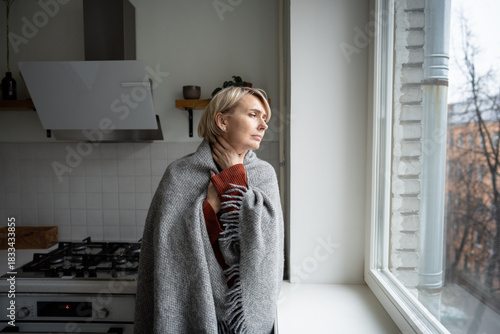 Middle aged woman covered in plaid stands near window touching throat with hand, getting sick having sore throat, seasonal symptoms of cold and flu caused by weather changes and heating problems.