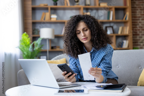 Woman holding a receipt and smartphone, comparing expenses while managing her budget at home, experiencing financial stress and worry over incoming bills