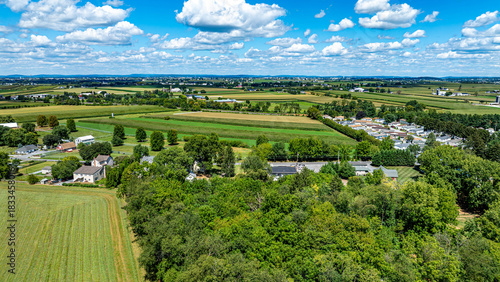 A vibrant rural landscape showcases green fields and distant farms under a blue sky filled with fluffy clouds. The scene is peaceful, emphasizing the beauty of nature and agriculture.