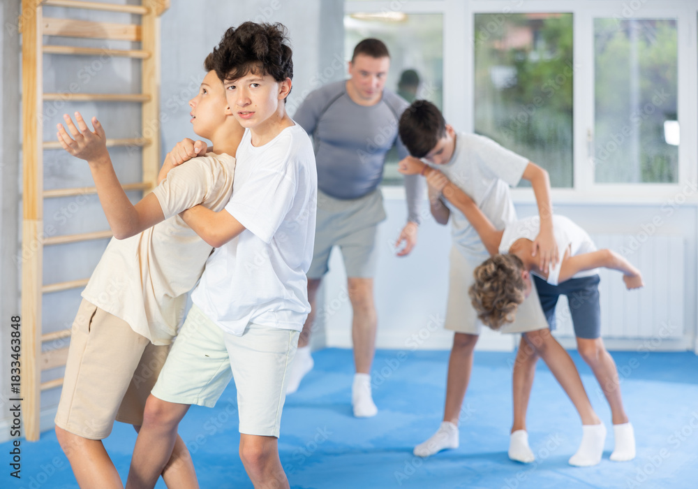 Obraz premium Determined teenagers sparring at self-defense training, trying to break free of back grab. Boys practicing basic techniques under guidance of experience instructor....