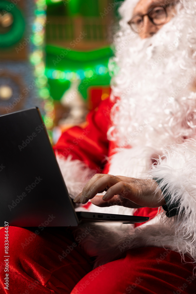 Fototapeta premium Santa using laptop to spread Christmas spirit virtually by sending cheerful greetings. Father Christmas prepares personalized messages for families using notebook, sharing positivity and joy