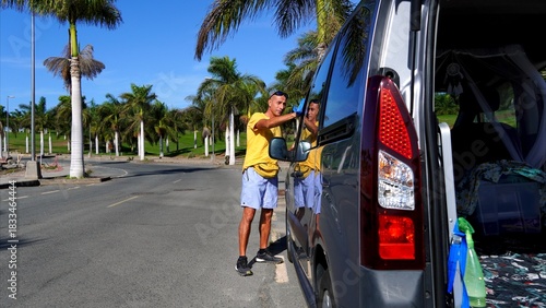 Mature man standing beside his camper van, meticulously cleaning the side mirror, representing car maintenance, personal care, and travel preparation in a tropical outdoor setting