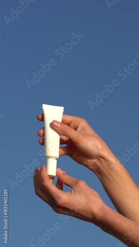 Vertical video of female hand holding white mockup tube of serum that squeeze and rubs into skin against clear, bright sky. Concept of anti-aging for daily routine, skin vitality, glow, and elasticity