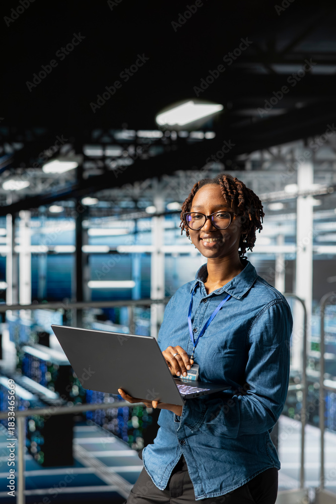 Fototapeta premium Portrait of black woman engineer solves maintenance tasks on laptop in high tech server hub, ensuring optimal performance and automation for software systems. Engineer reviewing diagnostics.