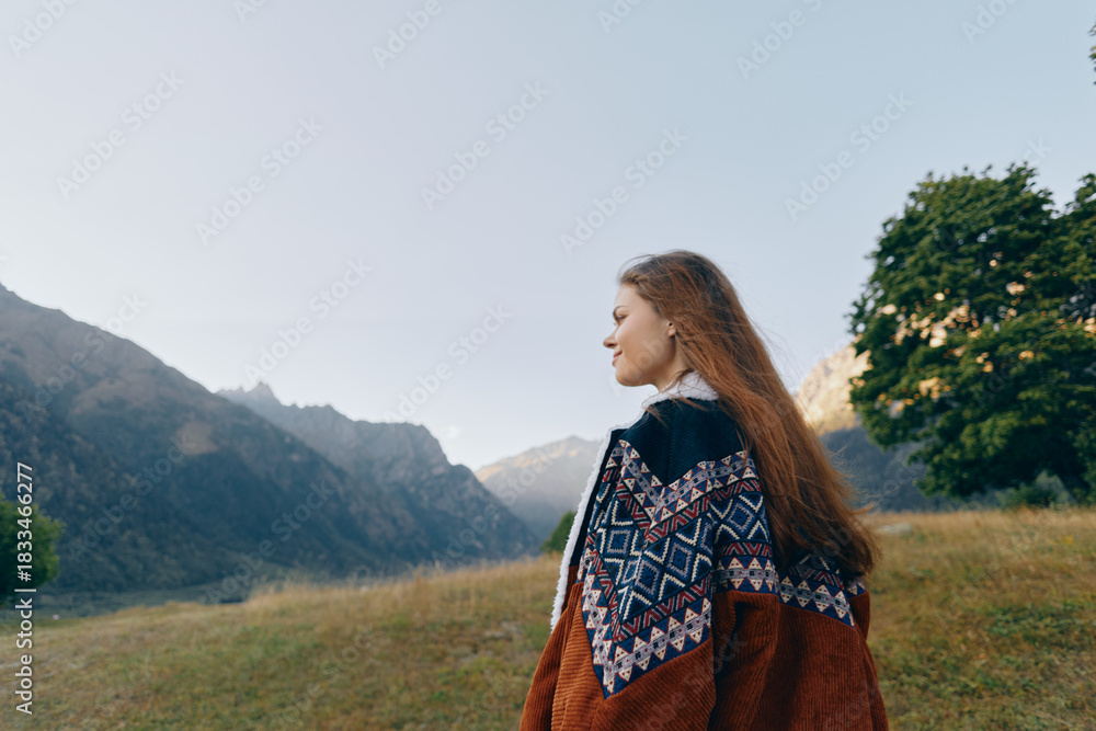 Fototapeta premium Woman portrait in sweater with long hair standing in mountain landscape meadow, looking sideways at distant peaks. Nature travel scene with trees, calm light, warm autumn mood and solitude.